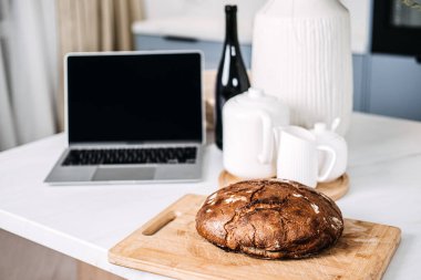 Dizüstü bilgisayarla mutfak masasında ev yapımı ekmek. Dizüstü bilgisayarın yanındaki kesme tahtasının üzerinde taze pişmiş ev yapımı ekmek, muhtemelen online bir yemek tarifini takip eden samimi bir ev yapımı sahneyi tasvir ediyor.