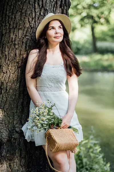 Eco-Friendly Lifestyle. Woman in Nature with Sustainable Fashion Choices. Outdoors woman embodying eco-friendly lifestyle with straw hat, dress, and basket, holding a bouquet of wildflowers