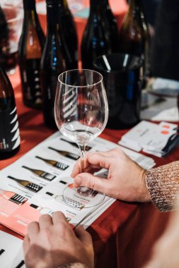 Katılımcı, profesyonel bir şarap tatma etkinliğinde şarap kadehinin üzerinde şarap bardağını inceler ve arka planda çeşitli şişeler bulunur..