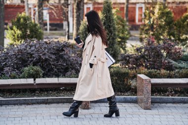 Elinde kahve fincanı ve dizüstü bilgisayarla parkta yürüyen bej ceketli meşgul bir kadın, doğa ve modern iş hayatının bir karışımı..