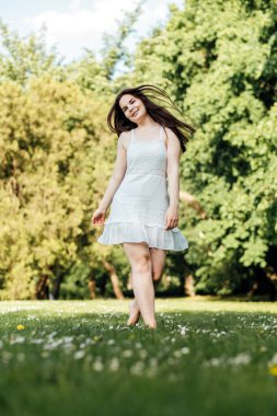 Beyaz bir yaz elbisesi içinde kaygısız bir kadın şehir parkındaki çimlerin üzerinde çıplak ayakla yürümekten hoşlanıyor..