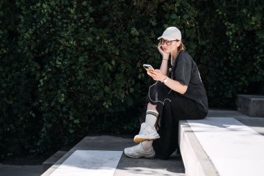 Rahat genç bir kadın telefonuyla müzik dinliyor, yeşil şehir yeşilliklerinin arka planında rahatça oturuyor..