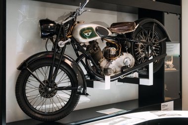 Parlatılmış metal motoru ve deri koltuğu olan klasik bir motosiklet, bir müze ortamında sergilenerek ulaşım geçmişini ön plana çıkarıyor..