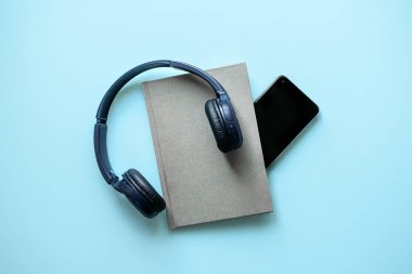 Bir çift kulaklık, yumuşak mavi arka planda bir akıllı telefonun yanında, modern sesli dinleme deneyimini tasvir eden bir kitabın üzerinde duruyor..