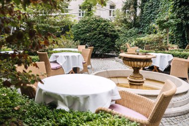 Hasır sandalyelerin, beyaz masa örtülerinin olduğu yuvarlak masaların ve yemyeşil bir çeşmenin bulunduğu zarif bir açık hava yemek alanı..