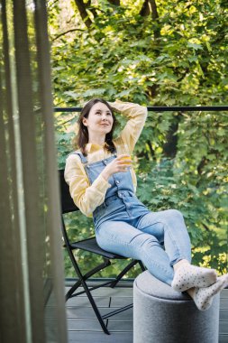 Genç bir kadın balkonda dinleniyor, meyve suyunu yudumluyor ve doğayla çevrili huzurlu sabahın tadını çıkarıyor. Görüntü, sabah ayinlerinin ve güne sakin başlamanın önemini vurguluyor..
