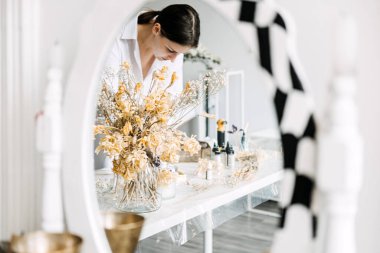 Sanat malzemeleri ve kurumuş çiçeklerle çevrili, aynaya yansıyan bir sanatçıyla sakin bir çalışma alanı. Yaratıcı uğraşlara girişmenin terapatik doğasını vurgular.