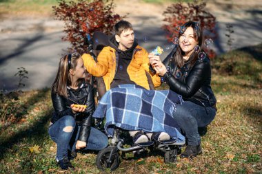 Bir anne ve kardeş, açık hava baloncuklarıyla tekerlekli sandalyedeki bir çocukla neşeli anlar yaratıyor. Görüntü, aile desteğini ve özel ihtiyaçları vurguluyor,