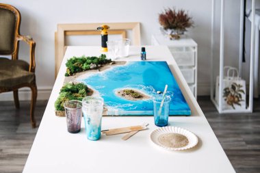 A resin art project featuring a coastal landscape with a beach and ocean, placed on a worktable in a home studio. The image highlights a creative DIY craft process with nature-inspired, handmade decor clipart