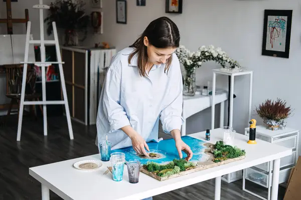 stock image A woman carefully crafts epoxy resin art, shaping an ocean scene with sand and greenery in a well-lit studio. The creative process highlights meticulous attention to detail, showcasing her artistic