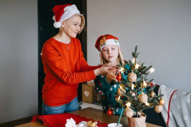 Anne ve kızının birlikte küçük bir Noel ağacını süslemesi, bayram bağlarının, aile rollerinin ve şenlik hazırlıklarının sevincinin vurgulanması.