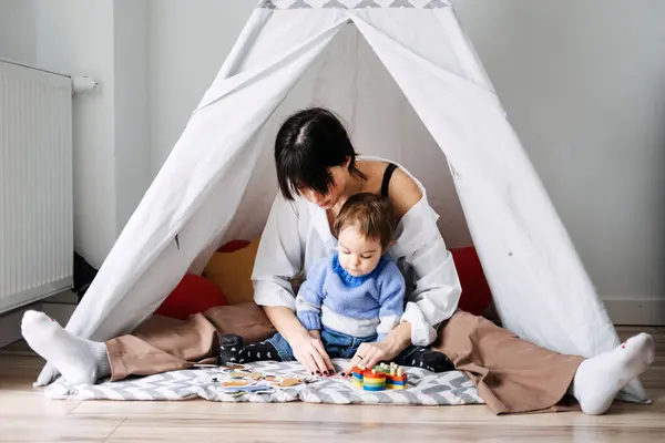 stock image A mother engages in play with her toddler inside a makeshift indoor tent, emphasizing the role of play in early childhood development and motor skills.