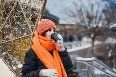 Şenlik ortamında turuncu fularlı ve şapkalı bir kadın kahve içiyor. Tatil modası, kentsel kış kutlamaları ve mevsimsel sıcaklık..