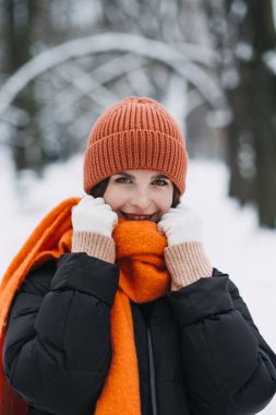 Woman in orange winter hat and scarf smiling in snow-covered park. Warm seasonal fashion, winter style trends, cozy outdoor clothing. Color Therapy in Seasonal Fashion. clipart