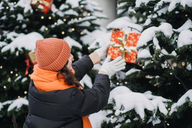 Karla kaplı bir Noel ağacını hediye kutularıyla süsleyen bir kadının arka görüntüsü. Tatil dekorları ve şenlikli kış manzaraları..