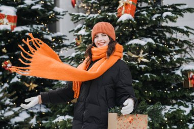 Parlak turuncu eşarplı bir kadın Noel ağaçlarının önünde hediye çantası tutuyor, neşeli tatil ruhunu kucaklıyor ve çevreci bir hediye veriyor. Tatil dekoru, kış neşesi