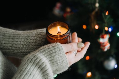 Elleri sıcak süveter içinde mum tutarken, Noel için dilek tutarken. Arka planda Noel ağacı olan ılık ve şenlikli bir tatil ortamı