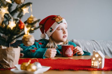 Noel Baba şapkalı genç bir kız, Noel süslemeleri ve tatil konforuyla çevrili evinde sıcak bir fincan kakaonun tadını çıkarıyor..