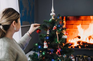 Bir kadın, Noel ağacını şöminesi olan rahat bir yazlık evde süslerle süslüyor. Sıcak mevsimlik dekorlu şenlikli ev tasarımı