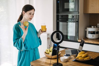 Sağlıklı yaşam tarzıyla sosyal medya içeriği yaratan genç bir kadın. Mutfakta meyve suyu var. Sağlıklı yaşam ve gıda vloglama kavramı.