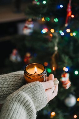 Elleri sıcak süveter içinde mum tutarken, Noel için dilek tutarken. Arka planda Noel ağacı olan ılık ve şenlikli bir tatil ortamı