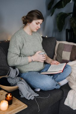 Kanepede sıcak içecekle kitap okuyan hamile bir kadın. Etrafı örgü malzemeleriyle çevrili. Hamilelik boyunca rahatlama, rahatlama ve kızlık zarı.