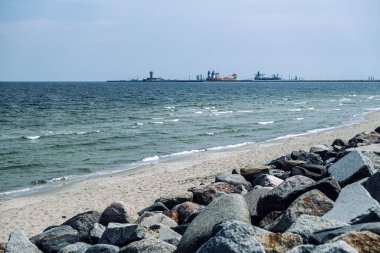 Rocky kıyı şeridi kumlu sahilde dalgalar ve endüstriyel gemilerin ve kontrol kulesinin uzaktan görüntüsü. Kıyı sahnesi. Gdansk, Polonya - 19 Mayıs 2024.