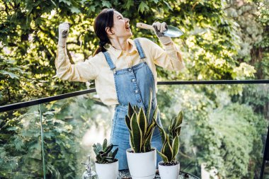 Elinde kürek tutan bir kadın, güneşli bir balkonda saksı bitkileriyle çevriliyken neşeli bir poz veriyor. Eğlence ve çevre bilinci olan bahçıvanlığı temsil eder