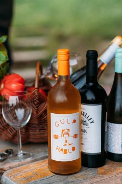 Assorted wine bottles with unique labels, a wicker basket, and glassware on a rustic wooden table in an outdoor setting clipart