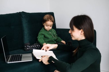 Bir anne, dizüstü bilgisayarı ve yanında yürümekte olan çocuğuyla koltukta çalışır. Evde iş ve çocuk bakımının dengelenmesinin zorluklarını vurgular..
