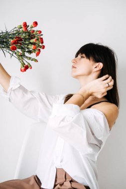 Kırmızı karanfilleri tutan bir kadın, yüzünü çiçeklerin arkasına gizliyor. Görsel hikaye anlatımı, çiçekli estetik ve minimalist kompozisyonu temsil eder, sosyal medya içeriği için mükemmeldir..