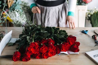 Çiçekçi buket hazırlığı sırasında kırmızı güllerin arkasında duruyor. Küçük çiçekçilik, kasıtlı tasarım ve zanaat odaklı girişimciliği temsil eder..
