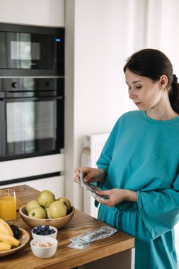 Mutfak tezgahının yanında meyve suyu ve meyve suyuyla Amerikan doları sayan genç bir kadın. Sağlık çağında bütçe belirleme, bilinçli harcamalar, finansal bakım, gıda maliyeti planlaması. Yüksek kalite fotoğraf