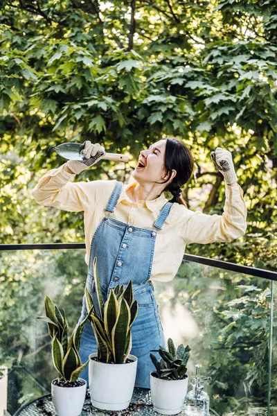 Genç bir kadın yemyeşil ağaçlarla çevrili bir balkonda saksı bitkileriyle neşeyle ilgileniyor. Duygusal destek tesisleri, yetiştirme alışkanlıkları, Z jenerasyonu hobileri, sürdürülebilir yaşam. Yüksek kalite fotoğraf