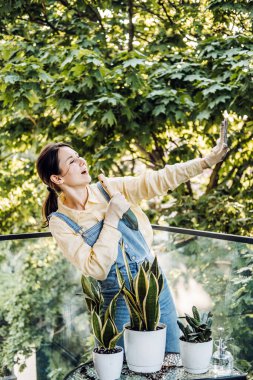 Genç Asyalı kadın balkon bitkileriyle ilgilenirken bir bahçe malasına şakıyarak şarkı söylüyor. Bitki terapisi, Z jenerasyonu kendini ifade etme, duygusal sağIık, sürdürülebilir hobiler. Yüksek kalite fotoğraf