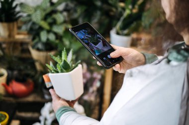 Bitkilerle dolu bir stüdyo ortamında beyaz orkideleri akıllı telefonuyla fotoğraflayan bir kadın. Çiçek girişimciliği, küçük işletme görselleri, çiçeklerle pazarlama, marka anlatımı. 