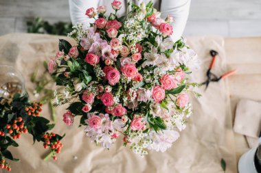 Üzerinde pembe güller, alstromerya ve çiçekçi masasında balmumu çiçekleri bulunan yemyeşil bir el yapımı buket. Lüks çiçekçilik, el yapımı buket, mevsimlik çiçekler, zarif çiçek düzenlemeleri.. 