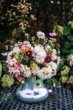 Mozaik bahçe sandalyeleri ile çevrili işlenmiş demir bir masaya yerleştirilmiş, gevşek sıralanmış bahçe çiçekleriyle dolu gümüş bir vazo taşar. Cottagecore estetiği, bahçe stili, çiçek bolluğu, yavaş yaşam.. 