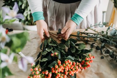 Çiçekçi, kraft kaplı bir masada portakal çilekleri ve okaliptüs dallarıyla bir buket hazırlıyor. Yavaş çiçekçilik ve botanik sanatları, dikkatli üretim, zanaatkar çiçek tasarımı, çiçeklerin sağlığı. 