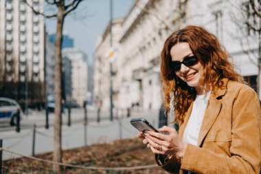 Güneşli bir şehir sokağında telefonu kullanarak gülümseyen içerik yaratıcısı. Mikro-etkiler, doğal bağlılık ve mobil hikaye anlatımı..