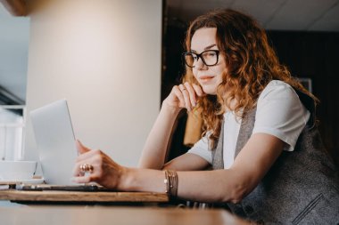Kıvırcık kızıl saçlı genç bir kadın laptopuyla hafif aydınlatılmış bir kafede konsantre oluyor. Odaklanmış üretkenlik, derin çalışma oturumları, dijital iş akışı, bilişsel katılım..