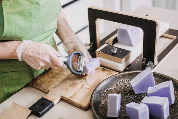 Ahşap bir yüzeyde el yapımı lavanta sabunlarının kenarlarını arıtmak için sebze soyacağı kullanan bir kadın. Sürdürülebilir cilt bakımı, ev bazlı iş modelleri, serbest iş, el işi ekonomisi. 