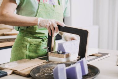 Büyük bir kalıp lavanta renkli sabunu dilimlemek için tel sabun kullanan yeşil önlüklü biri. Sanatsal sabun yapımı, küçük seri üretim, çevre dostu cilt bakımı, el yapımı ürün ekonomisi. 