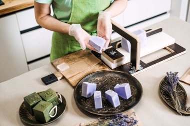 Büyük bir kalıp lavanta renkli sabunu dilimlemek için tel sabun kullanan yeşil önlüklü biri. Sanatsal sabun yapımı, küçük seri üretim, çevre dostu cilt bakımı, el yapımı ürün ekonomisi. 