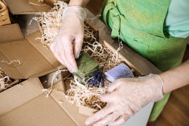 Eldivenli eller, kraft ambalajında kurumuş çiçeklerle yeşil ve lavanta sabunları düzenler. El yapımı iş, çevre dostu ambalaj, doğal cilt bakımı, sanatsal sunum. Yüksek kalite fotoğraf