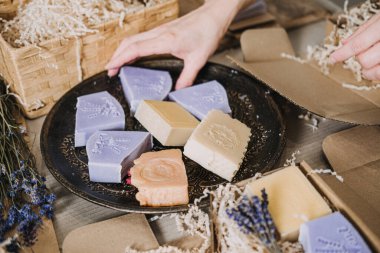 Eller, kraft ambalajı ve kurutulmuş lavanta ile siyah bir tabakta renkli el yapımı sabunlar düzenler. Sürdürülebilir sabun, plastiksiz yaşam tarzı, ekolojik bilinçli ürünler, yeşil ambalaj. Yüksek kalite fotoğraf