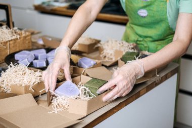 Kişi, lavanta ve zeytinyağı sabunlarını bir çalışma masasında öğütülmüş dolgu malzemesi ile birlikte kraft kutularına yerleştirir. Yeşil iş, sürdürülebilir ambalaj, sabun imalatı, çevre dostu uygulamalar. kalite fotoğraf