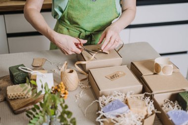 Kişi, Kraft sabun kutularını dilimlenmiş dolgu malzemesi ve el yapımı ürün etiketleriyle bir masaya yerleştirir. El yapımı hareket, analog yaratıcılık, DIY ambalajlama, insan eli değmesi. Yüksek kalite fotoğraf