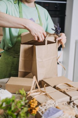 El yapımı sabunlu hediye kutuları mutfak masasına konur. Kişi de kese kağıdını bağlar. DIY iş görselleri, el yapımı ürün ambalajları, girişimci hayatı, el işi fuarı hazırlıkları. Yüksek kalite fotoğraf