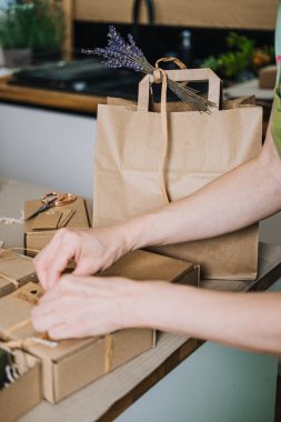 Kişi, kraft kutularını mutfak tezgahına, lavanta desenli bir kese kağıdının yanına iple bağlar. Çevre dostu ambalaj, plastiksiz hediye paketi, sürdürülebilir sunum, bağımsız nakliye. 